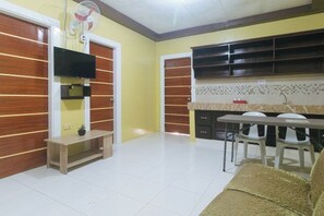 A living room with a sofa, a TV, a fan, a small table, and a kitchen area with a counter and chairs. - RedDoorz @ Queens Hometel (Tagum)