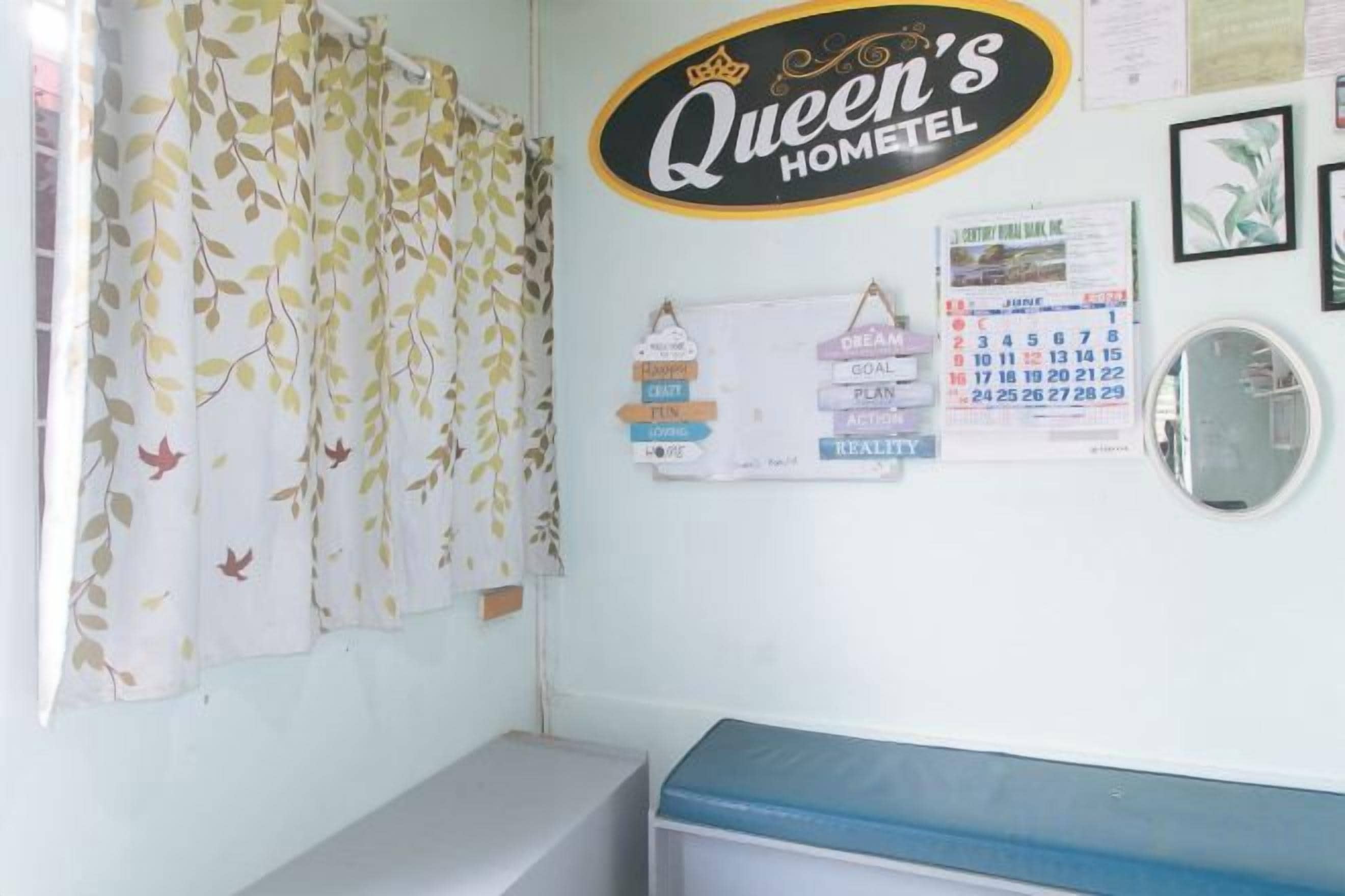 A hotel room with a bed, a small desk, a calendar, and a wall with a sign that reads 'Queen's Home'.