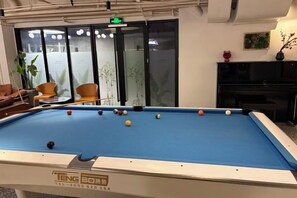 Une table de billard avec des billes, un canapé, des fauteuils, un téléviseur et un piano dans une pièce dont un mur est en verre.