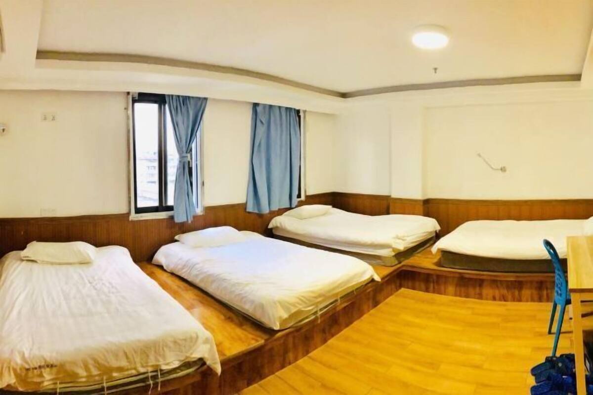 Een hotelkamer met drie bedden, geordend in een U-vorm, een houten vloer en een raam met gordijnen.