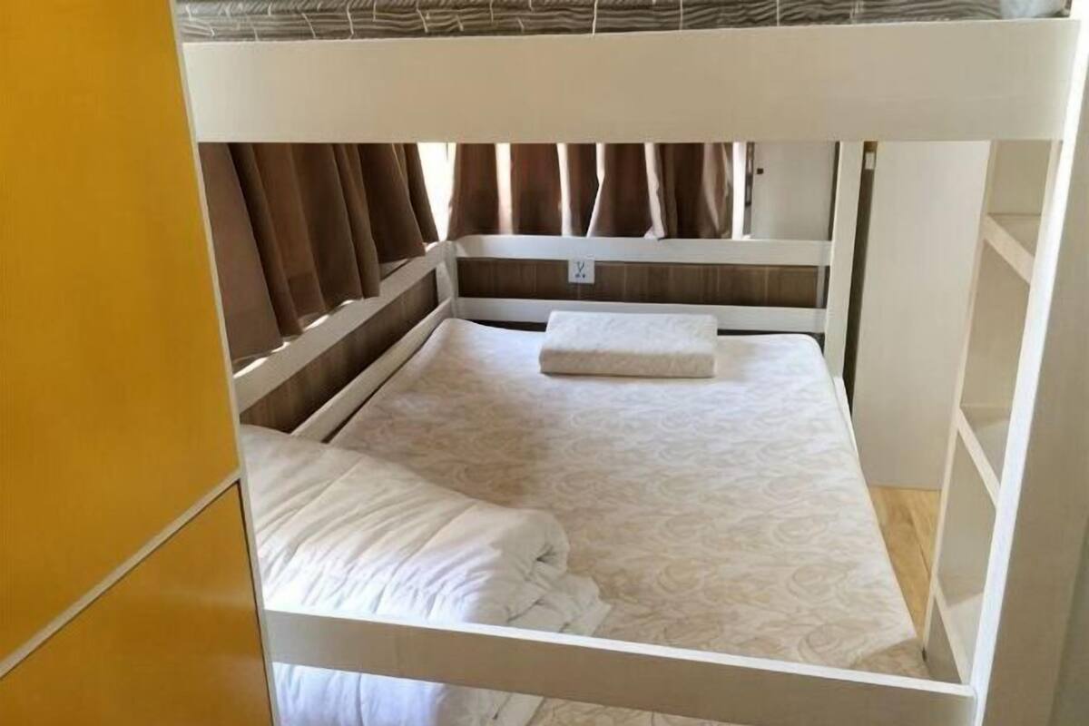 Een stapelbed met matras en kussens, een raam met gordijnen, en een lichtschakelaar aan de muur.