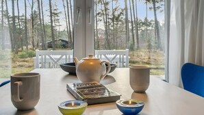 Una mesa con una tetera, tazas, un libro y dos velas en una habitación con vistas a árboles a través de una ventana.