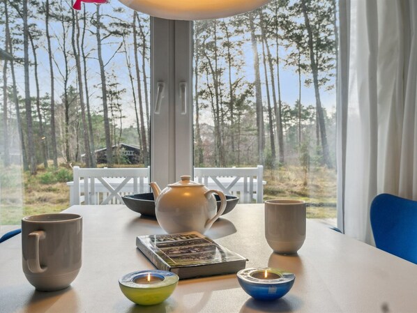 A table with a teapot, cups, a book, and two candles in a room with a view of trees through a window.