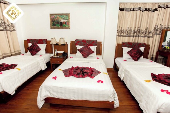 A hotel room with three single beds, each with a wooden headboard, white bedding, and red decorative pillows. There is a nightstand with a lamp and a phone, a painting on the wall, and a window with curtains.