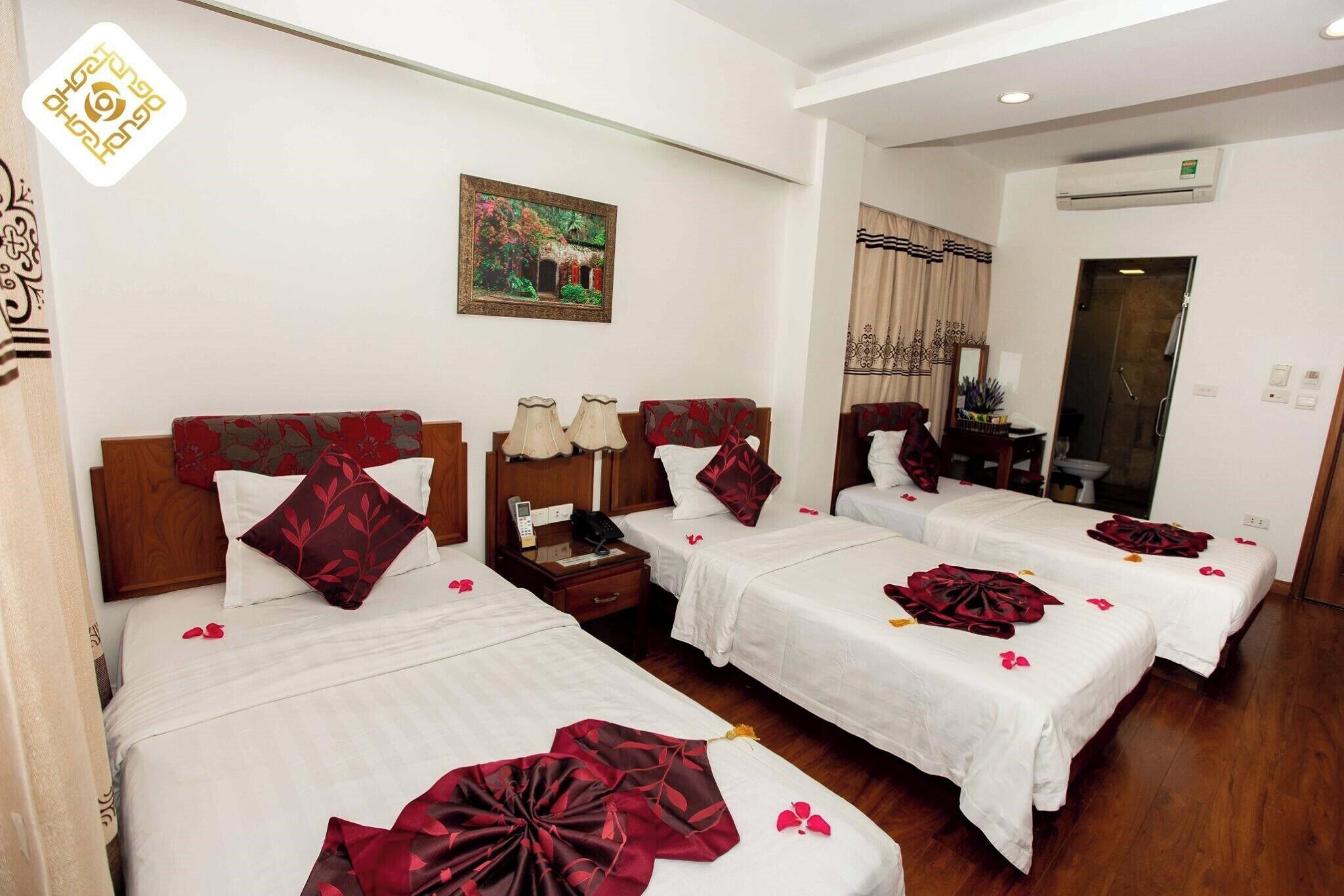 A hotel room with four single beds, each with a white bedspread and red decorative pillows. There is a painting on the wall above the beds, a wooden headboard, and a small table with a lamp between the beds.