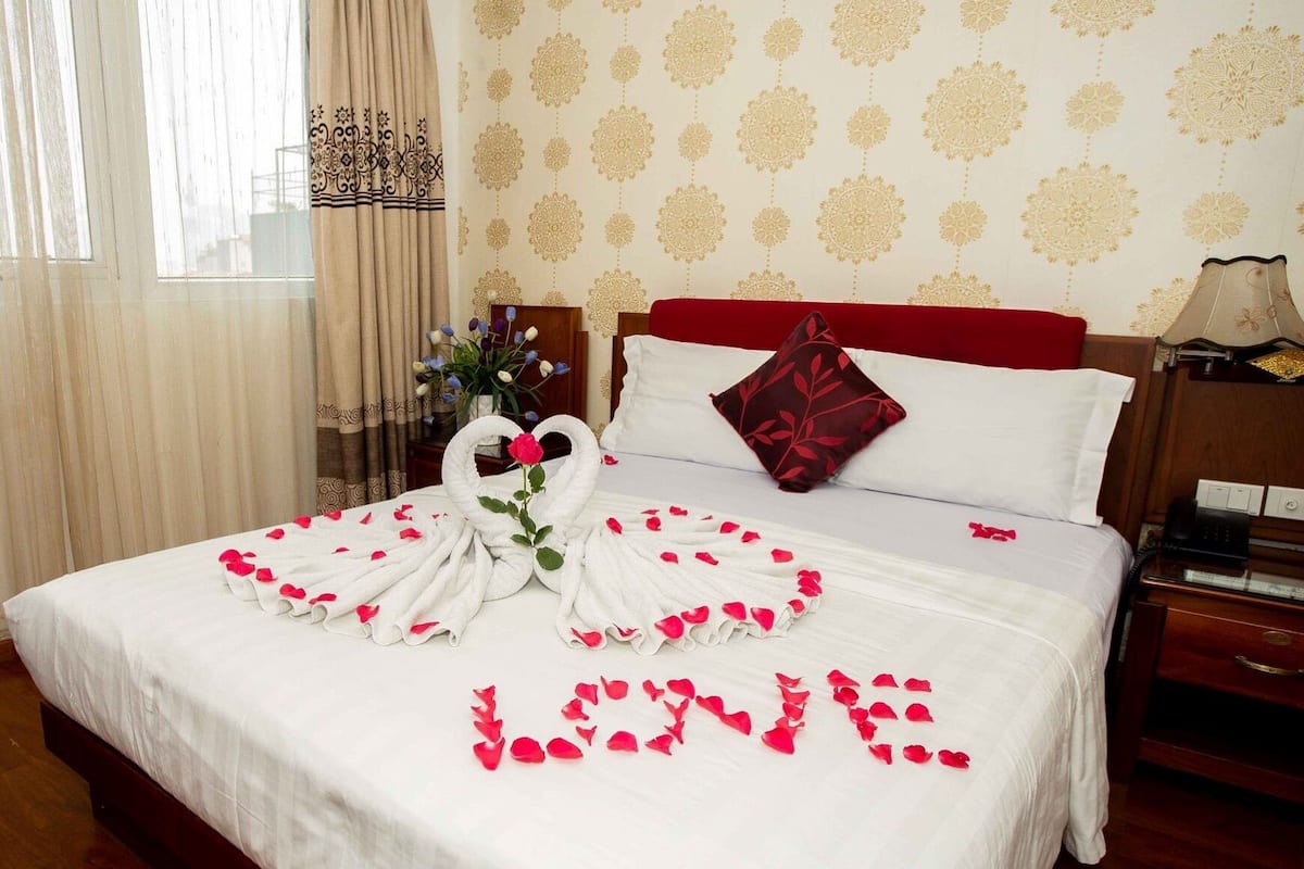 Habitació d'hotel amb un llit, un coixí vermell, un vas amb flors i una paret amb un dibuix floral.