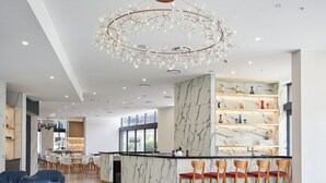 Un hall d’hôtel moderne avec un comptoir de réception en marbre, un coin bar avec des chaises en bois, un lustre et une moquette à motifs.