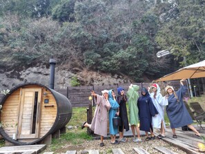 A group of people posing in front of a wooden structure with a barrel, a wooden fence, and trees in the background. - Tsuzuya Village, a guesthouse with a full-fledged private sauna (Minokamo)
