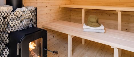 A wooden cabin interior with a wood-burning stove, benches, and a stone fireplace.