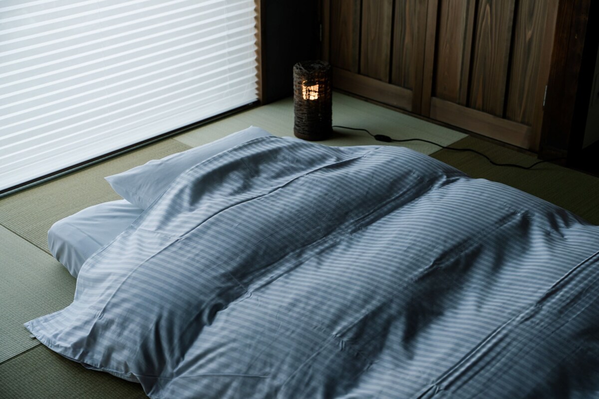 A room with tatami flooring, a bed with a striped bedding, a wall-mounted heater, and a window with blinds.