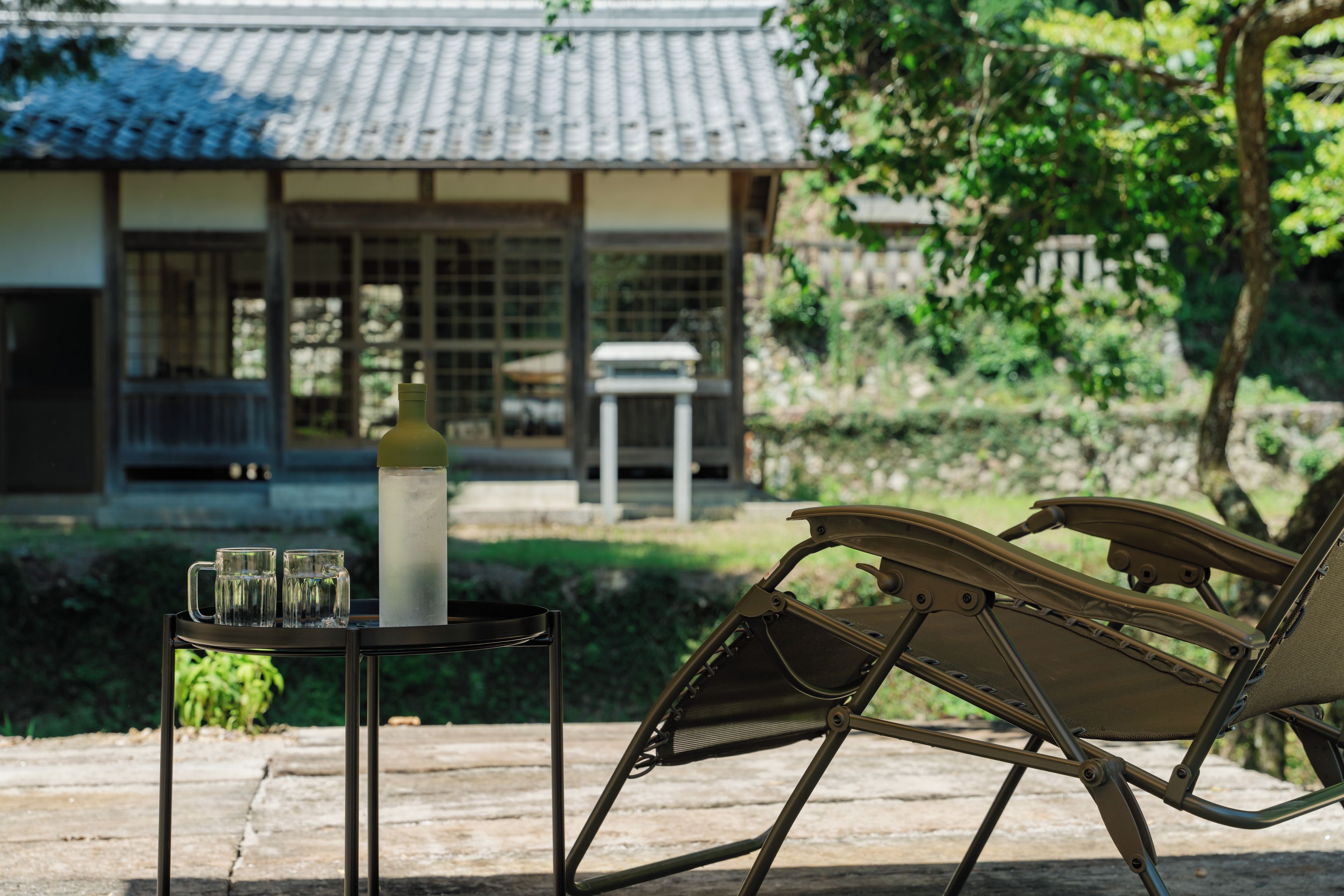 A traditional Japanese-style building with a tiled roof, a garden with a stone pathway, and a lounge chair with a metal frame.