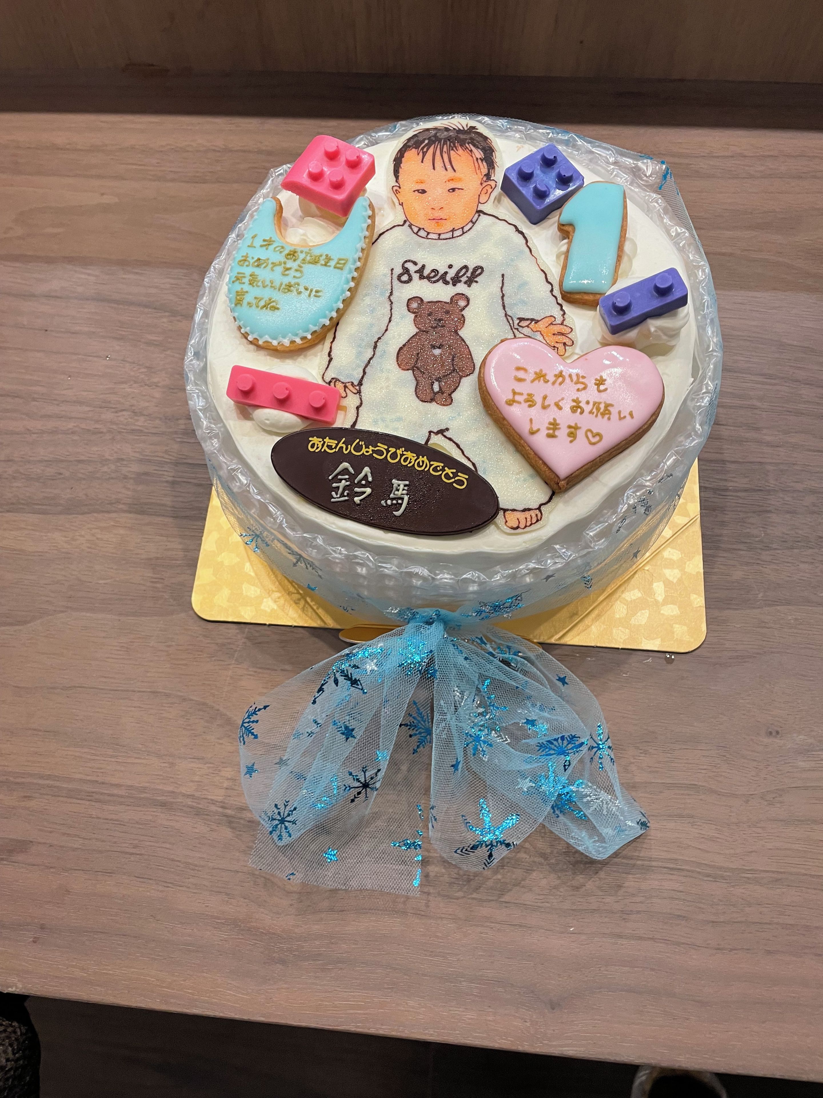 Un pastel redondo con un osito de peluche y decoraciones con forma de corazón, envuelto en plástico transparente y sobre un pedestal dorado.