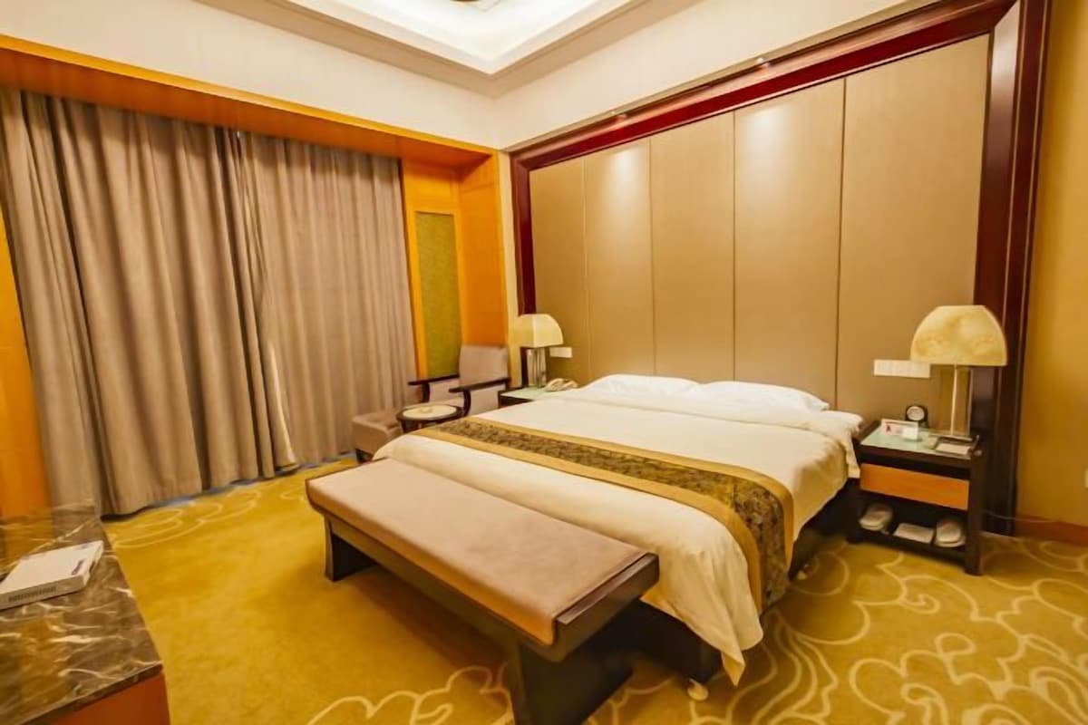 A hotel room with a bed, bedside tables, a bench, a desk, and a large window with curtains.