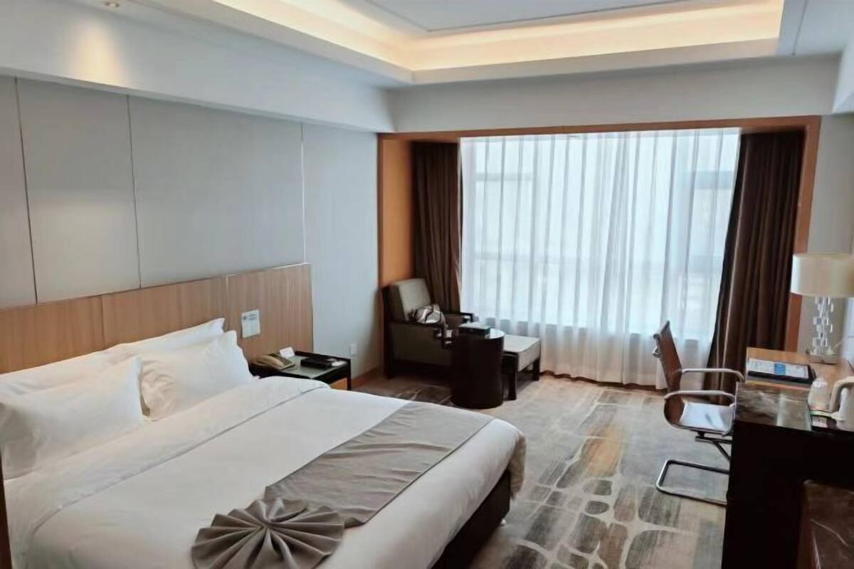 Een hotelkamer met een groot bed, een bureau, een stoel, een klein tafeltje en een raam met gordijnen.