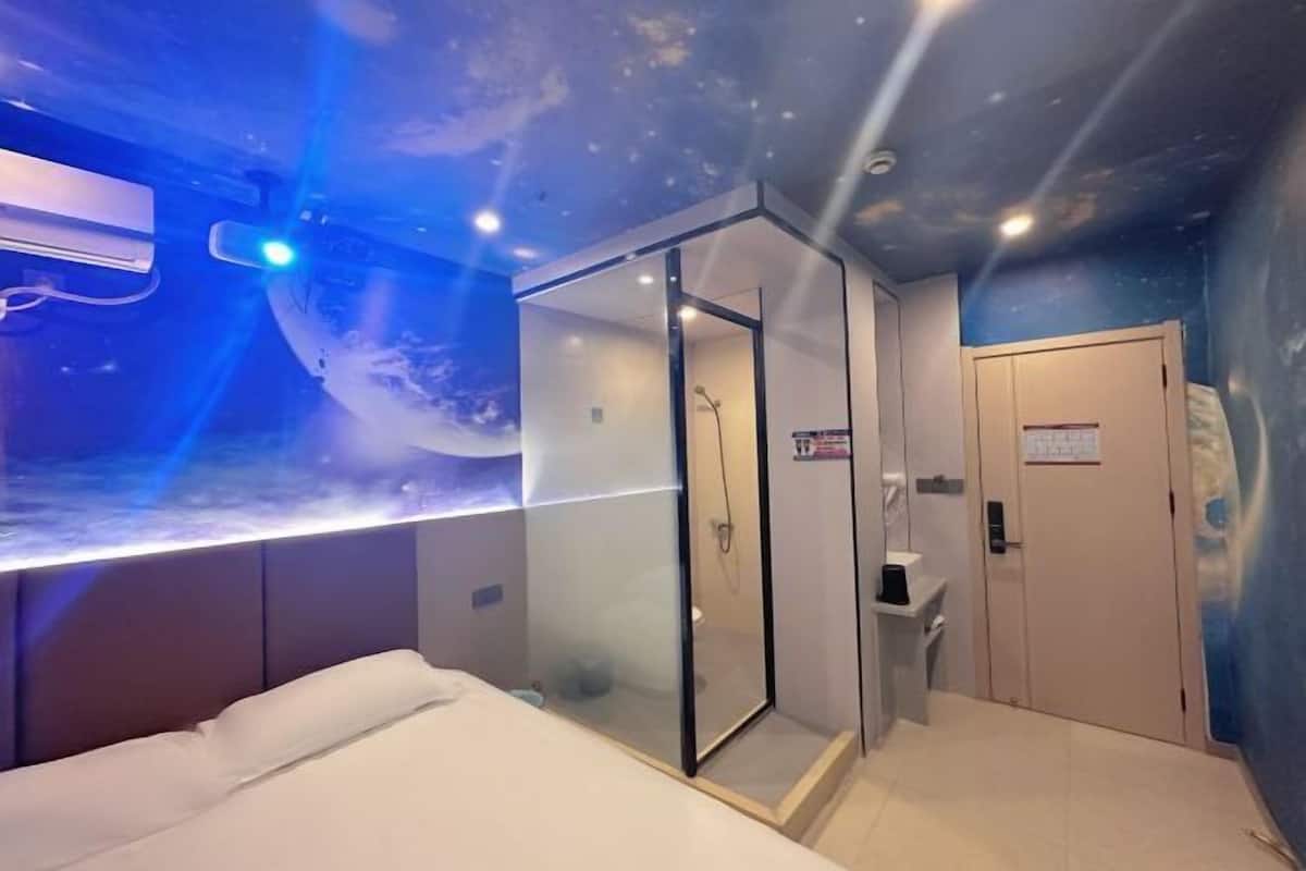 A room with a bed, a glass-enclosed shower, a door, and a wall with a space-themed mural.