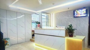 A hotel reception area with a marble wall, a reception desk with a yellow accent, a television mounted on the wall, and a potted plant on the desk. - Ningbo Jinhui Business Hotel (Ningbo)