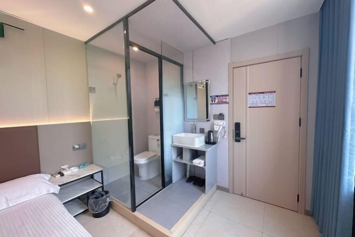 A modern hotel room with a bed, a glass-enclosed shower, a sink, and a door with a sign.