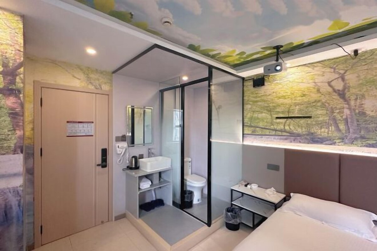 A modern hotel room with a glass-enclosed shower, a large bed, a bedside table, and a wall-mounted projector.