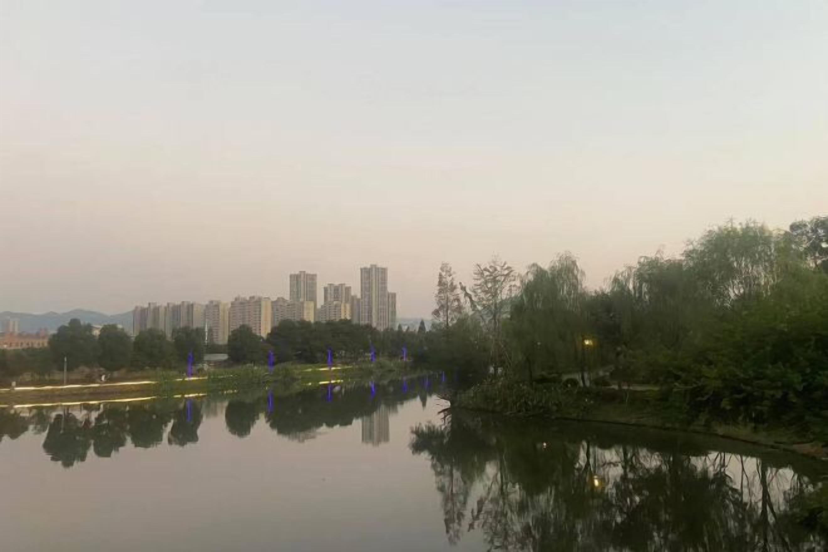 Un lago tranquilo con reflejos de árboles y edificios, rodeado de vegetación y algunas estructuras iluminadas.