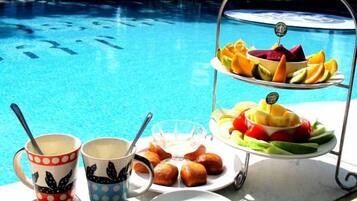 A poolside table with a fruit platter, two mugs, and a plate of pastries.