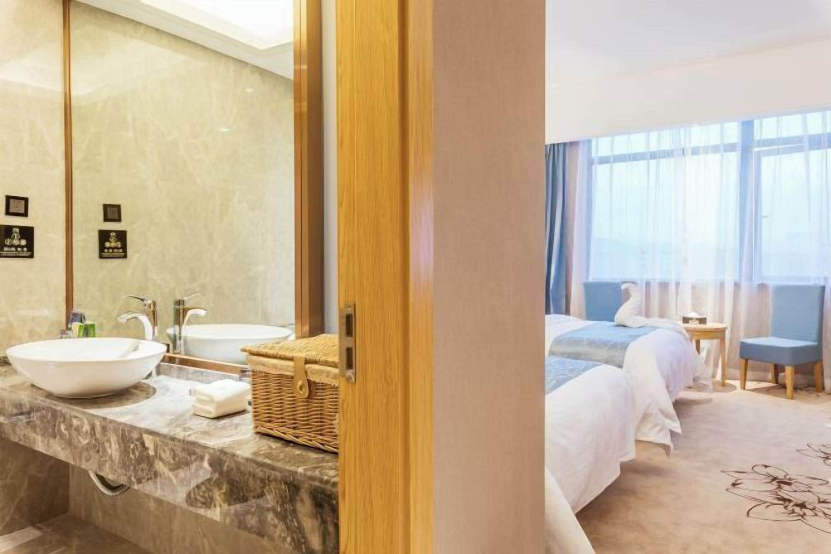 A hotel room with a marble bathroom vanity, two sinks, a large mirror, and a bed with a blue headboard.