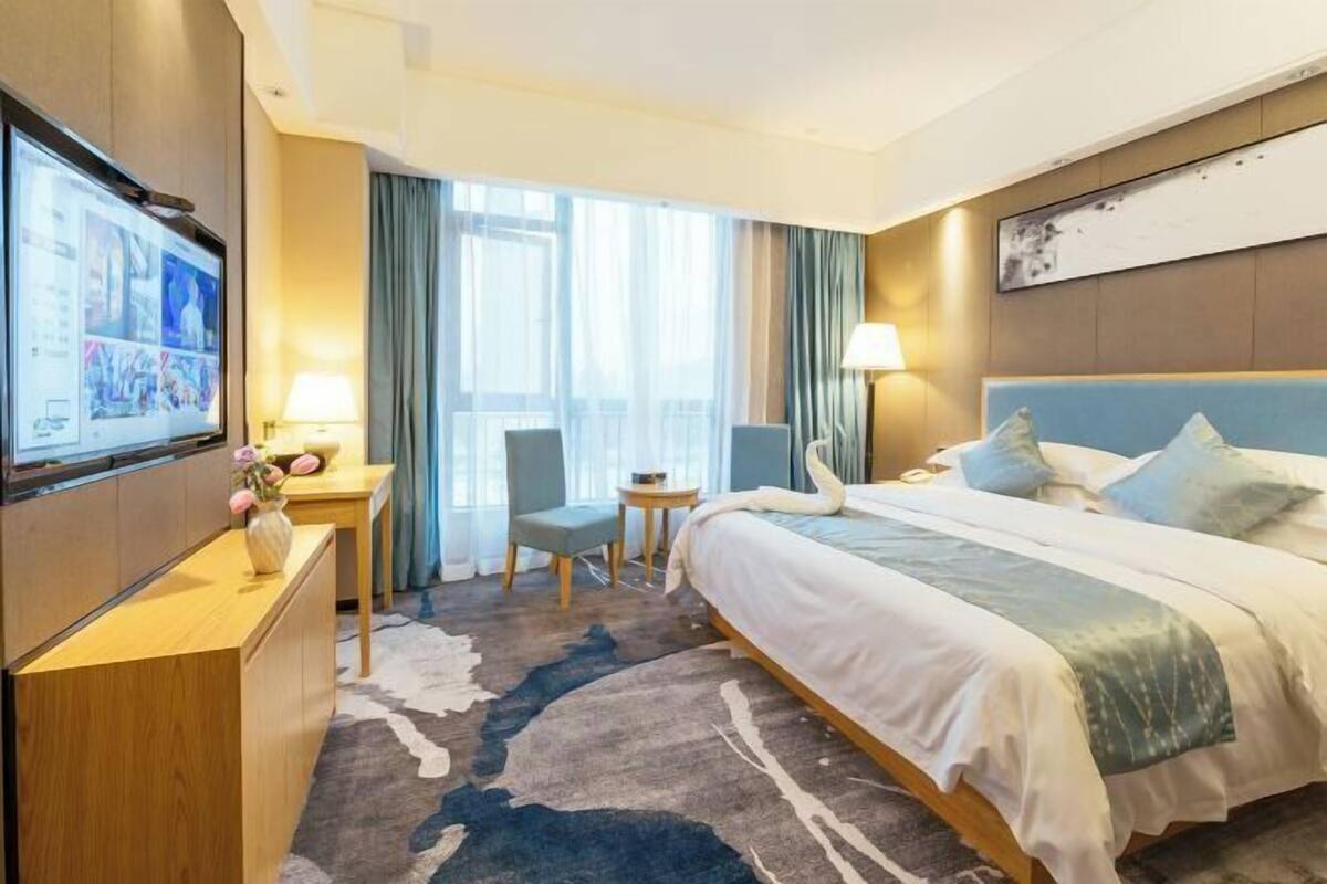 Une chambre d’hôtel avec un lit, un bureau avec une lampe, un téléviseur fixé au mur et une fenêtre avec des rideaux.