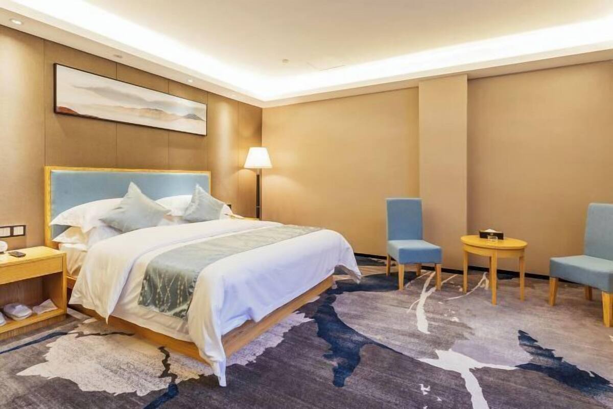 Une chambre d’hôtel avec un grand lit, deux chaises, une petite table, une lampe et un tableau au mur.
