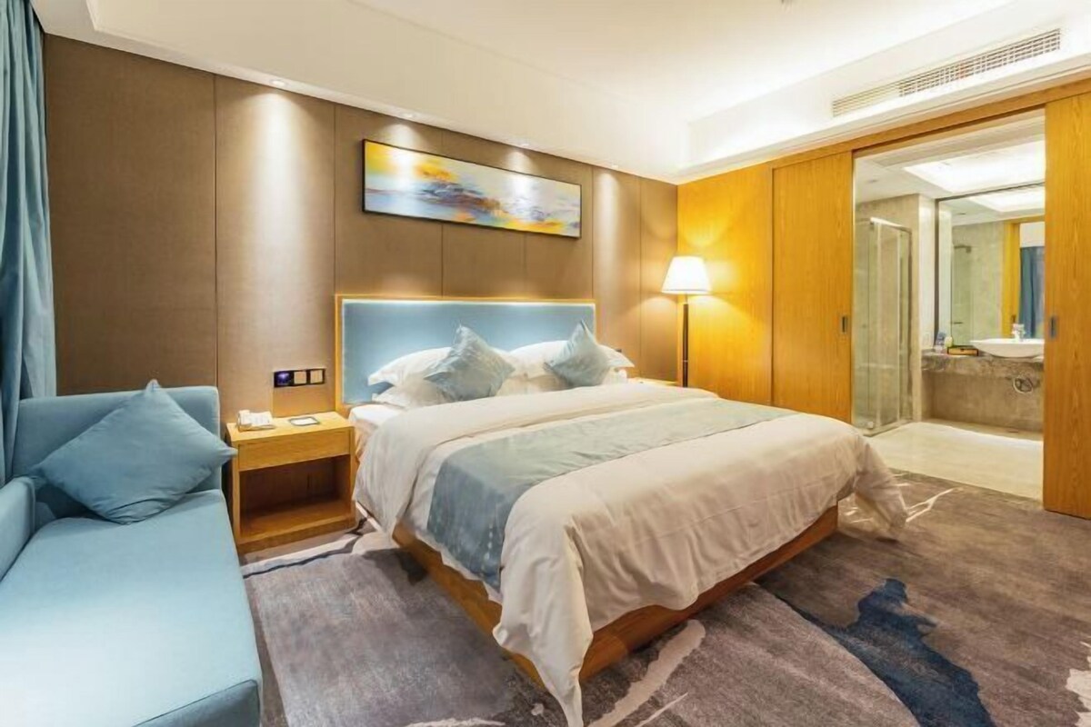 A hotel room with a bed, a bedside table, a lamp, a sofa, and a bathroom visible through an open door.