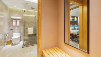 A modern bathroom with a glass shower enclosure, a wooden bench, and a large mirror reflecting a bedroom with a bed and a nightstand.