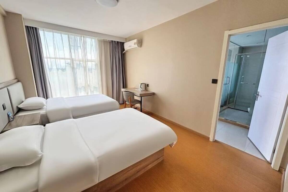A hotel room with two beds, a desk, a chair, a window with curtains, and a door leading to a bathroom.