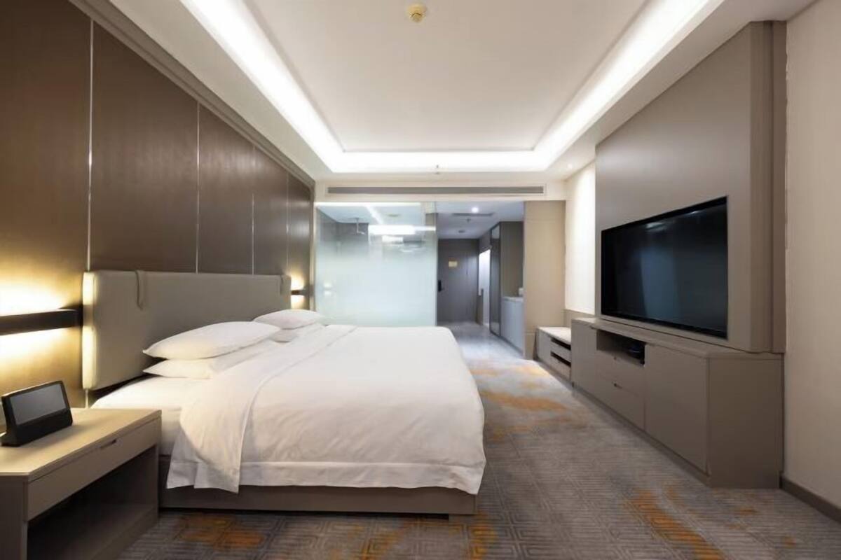 Ein modernes Hotelzimmer mit einem großen Bett, Nachttischen, einem Flachbildfernseher und Blick in die Hotellobby.
