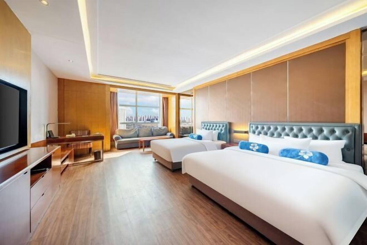 Ein Hotelzimmer mit zwei Betten, einem Fernseher, einer Sitzecke und einem großen Fenster mit Blick auf die Stadt.