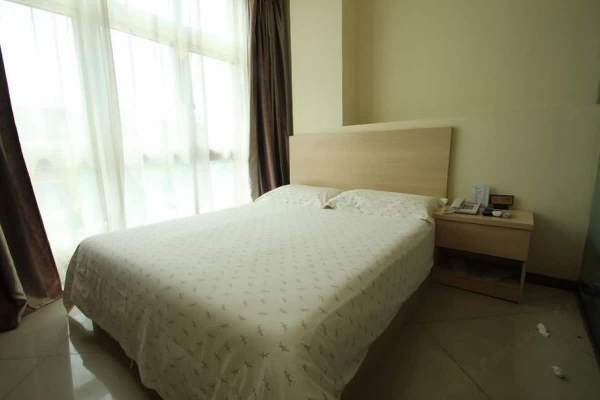 A neatly made bed with a patterned bedspread, a bedside table with a phone and a small item, and a window with curtains.