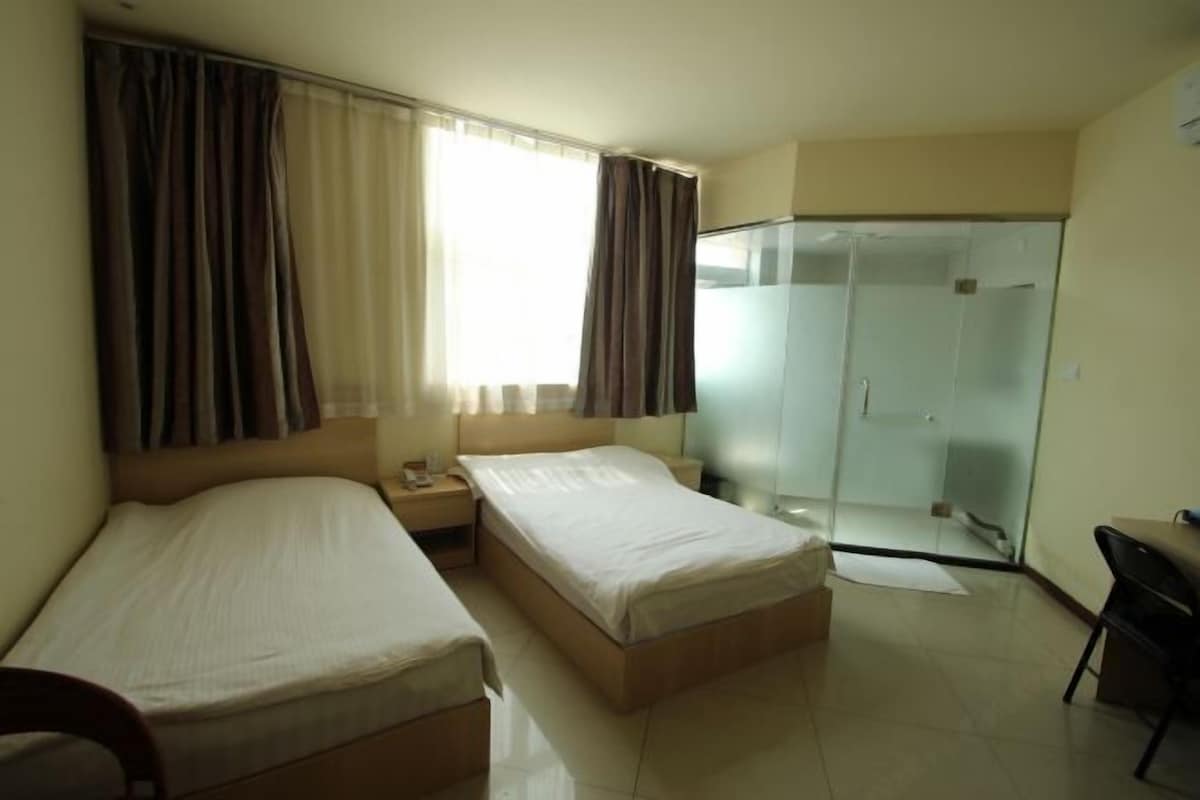 A hotel room with two beds, a desk, a chair, a shower, and a glass-enclosed bathroom.