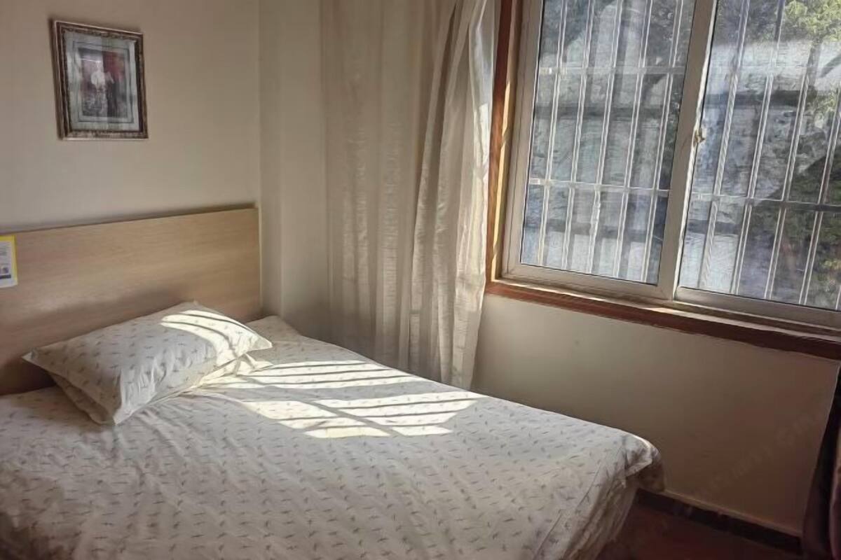 A single bed with a headboard, a window with vertical blinds, and a framed picture on the wall.