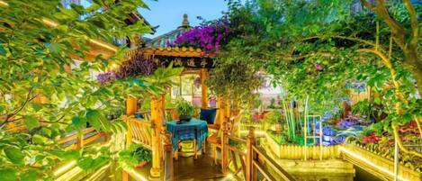 A wooden pathway leading to a covered seating area with a table and chairs, surrounded by lush greenery and a decorative archway.