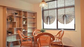 A room with a dining table, four wicker chairs, a wooden bookshelf, and a large window with a fan.