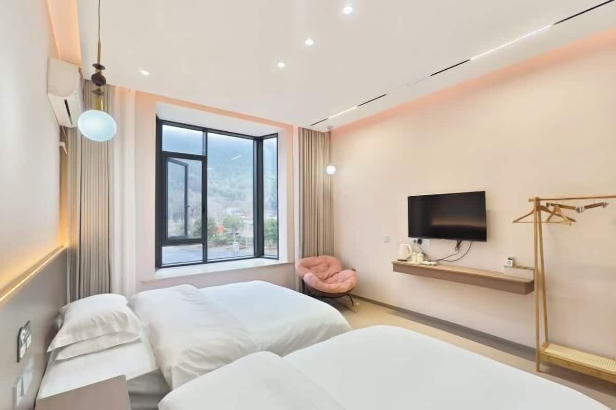 Une chambre d’hôtel moderne avec deux lits, une grande fenêtre, un téléviseur à écran plat, une chaise et une étagère en bois.