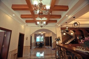 Un vaste hall d’entrée avec un plafond haut, des poutres en bois et un lustre. On aperçoit à l’arrière-plan un coin bar avec des tabourets et un comptoir.