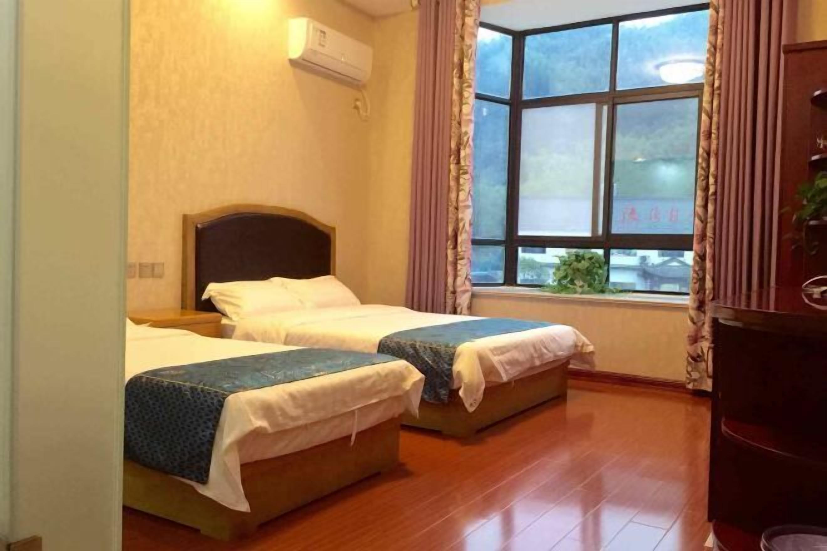 Habitación de hotel con dos camas individuales, suelo de madera, ventana con cortinas y un equipo de aire acondicionado en la pared.