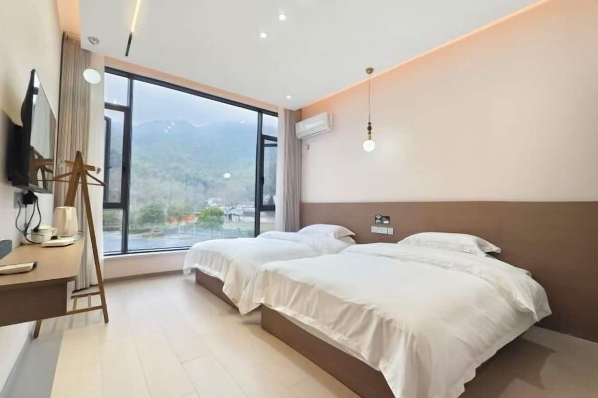 A modern hotel room with two beds, a desk, a chair, and a large window offering a mountain view.