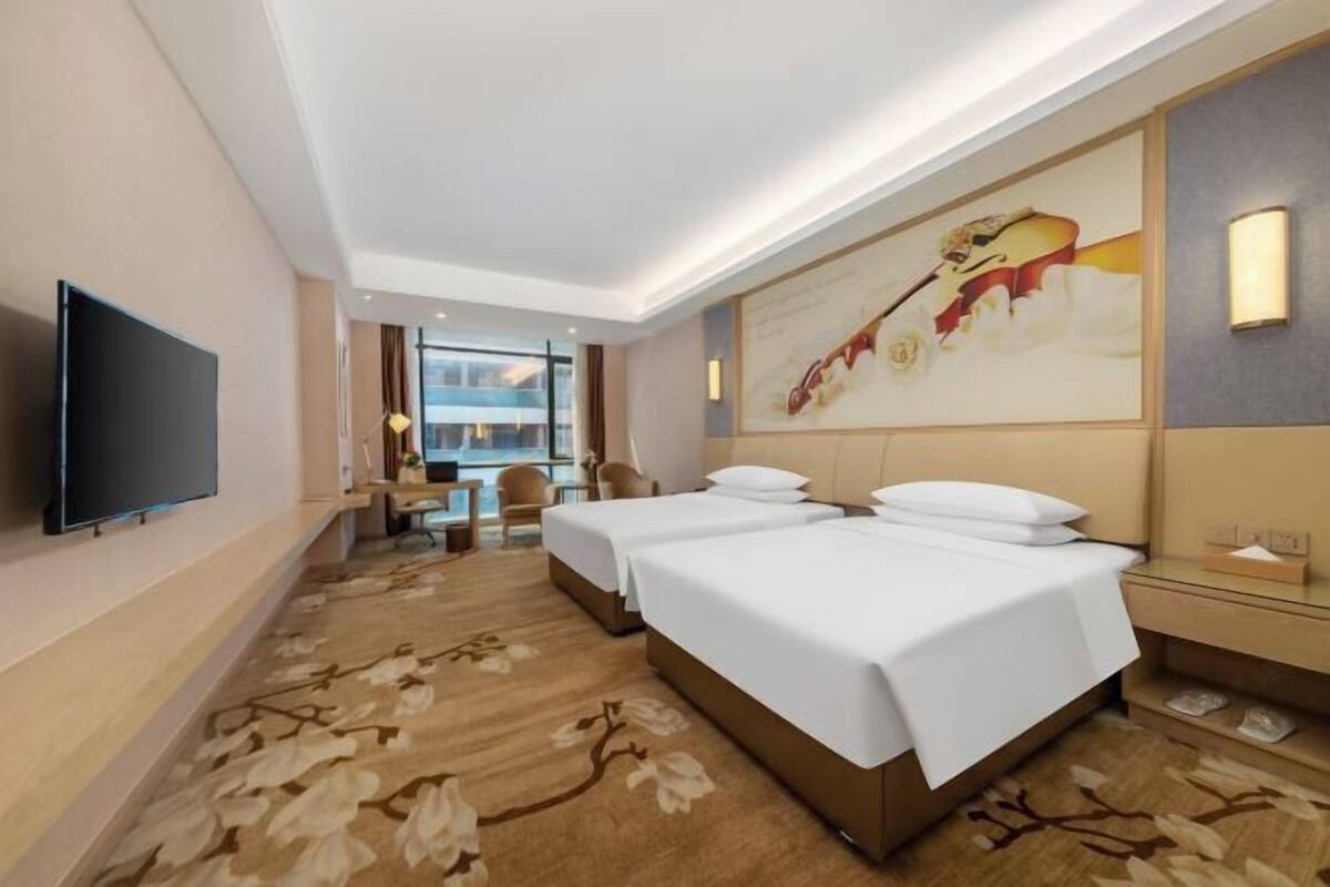 A hotel room with two beds, a flat-screen TV, a desk with a chair, a large window, and a painting on the wall.
