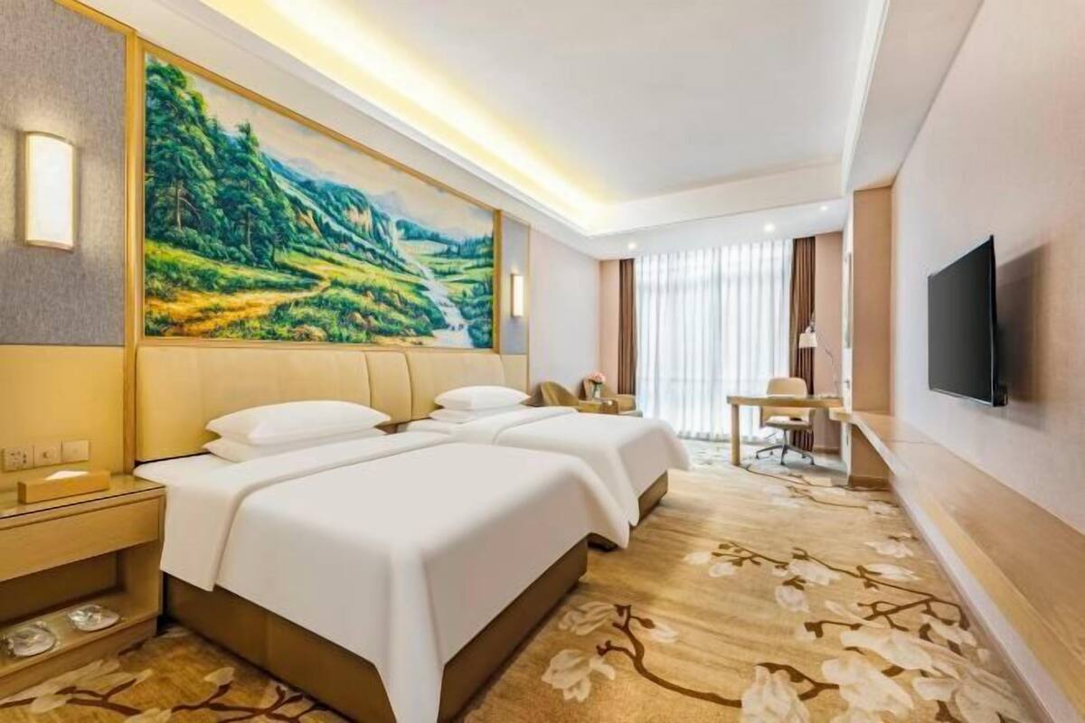 A hotel room with two beds, a desk, a TV, and a painting of a landscape.