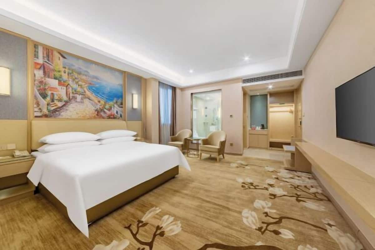 A hotel room with a bed, a desk, a chair, a painting on the wall, and a TV on the wall.