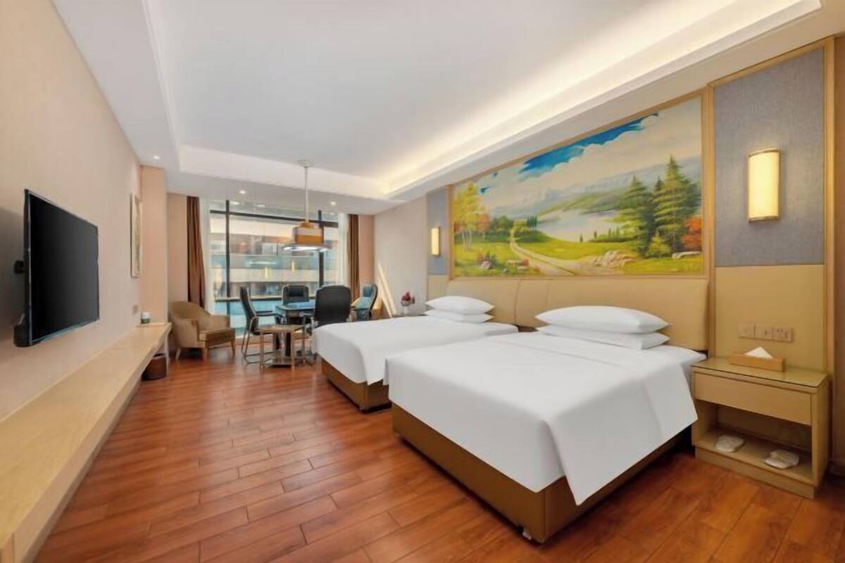 A hotel room with two beds, a desk, a chair, a TV, and a painting on the wall.