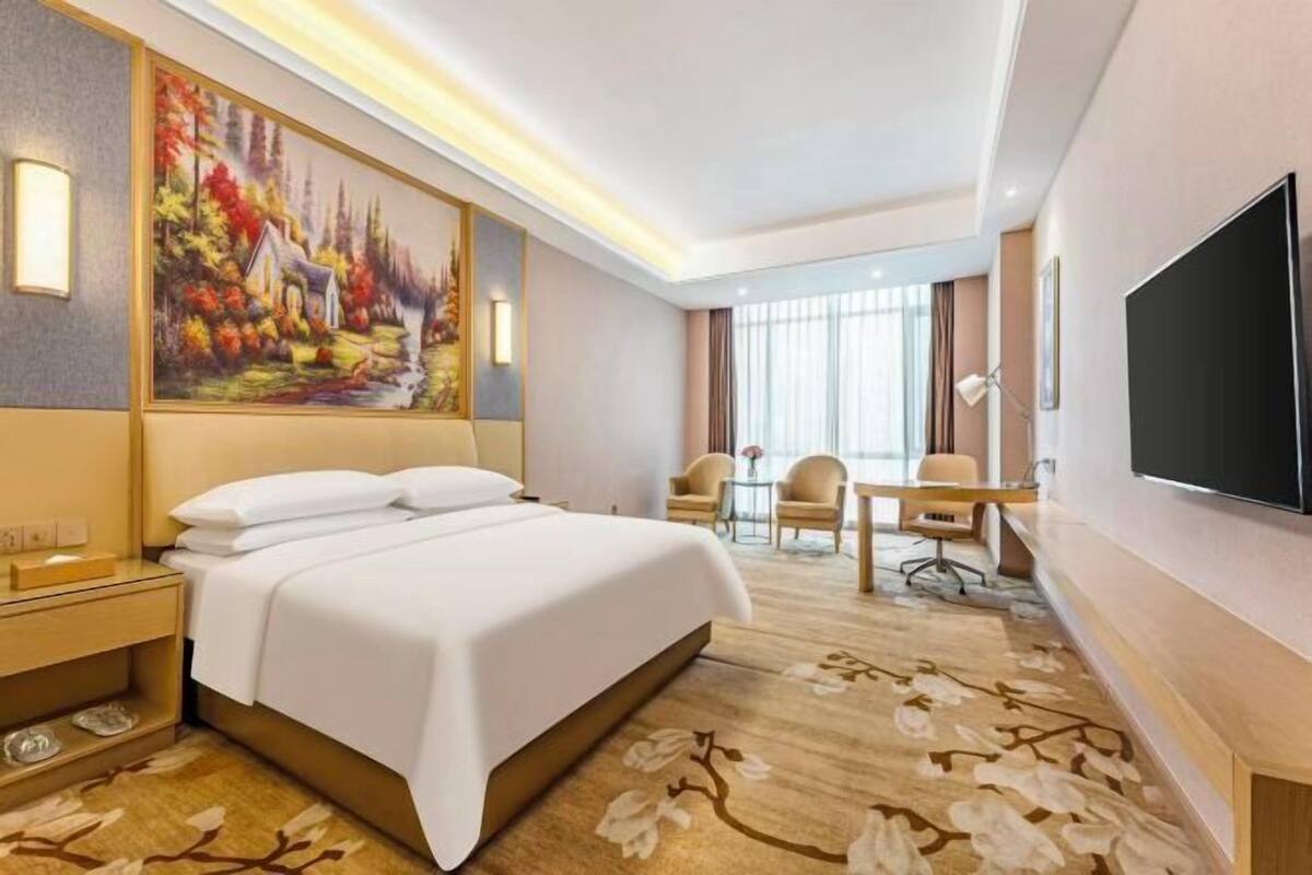 A hotel room with a large bed, a desk with two chairs, a television, and a painting on the wall.