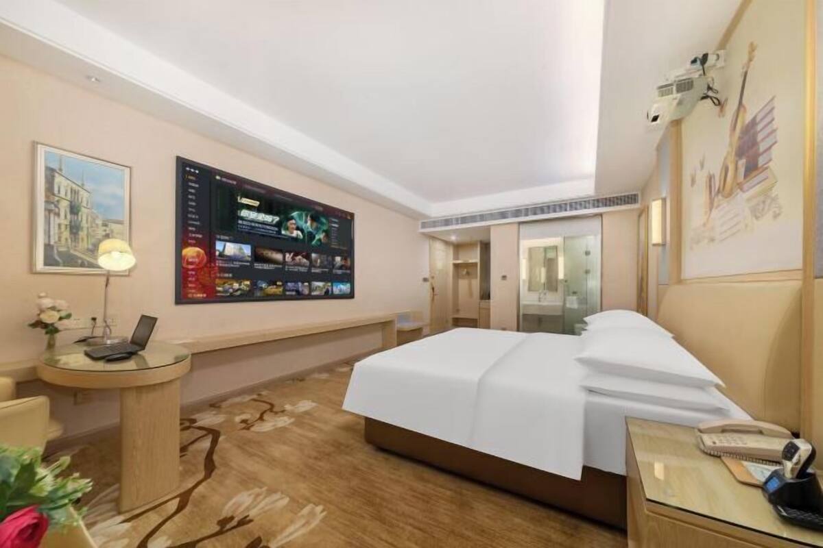 A hotel room with a bed, a desk with a laptop, a TV mounted on the wall, and a phone on the bedside table.