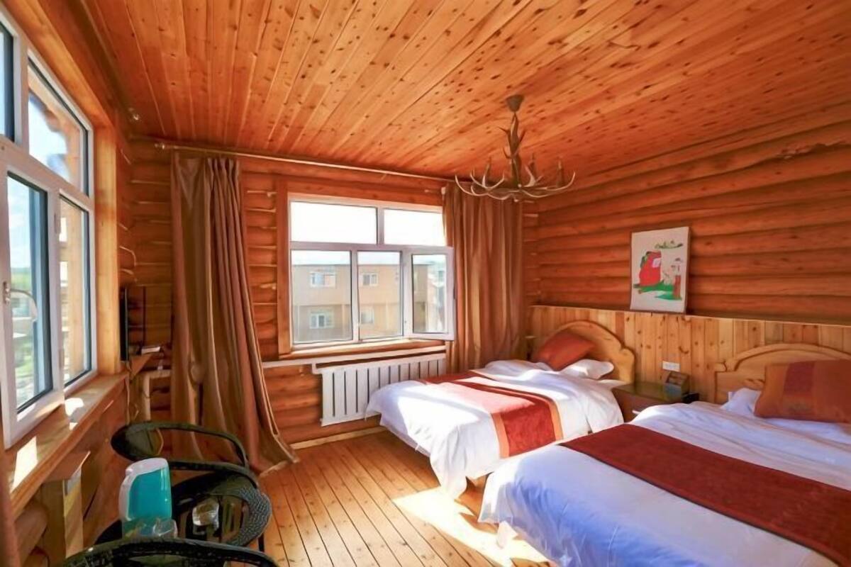 A wooden cabin bedroom with two beds, a chandelier, a window with curtains, and a radiator.