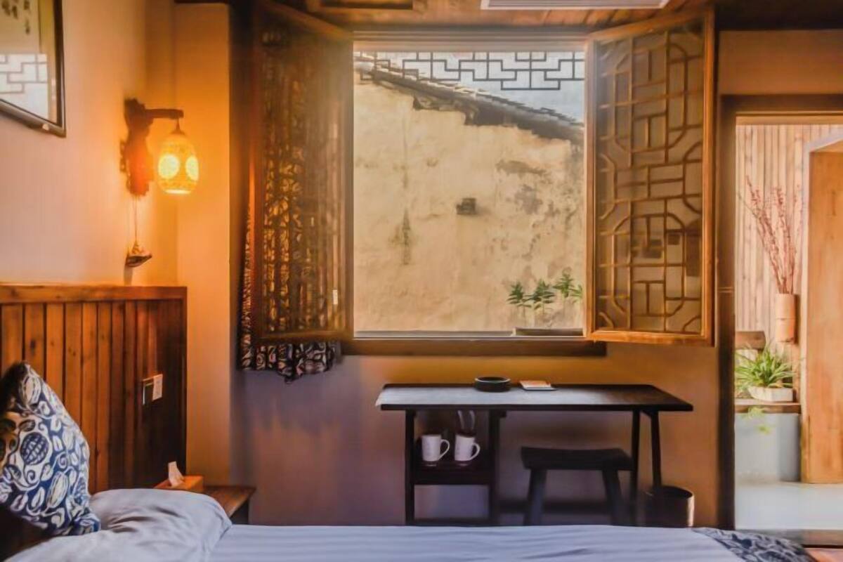A room with a bed, a table with a chair, a window with a view, and a wall-mounted light fixture.
