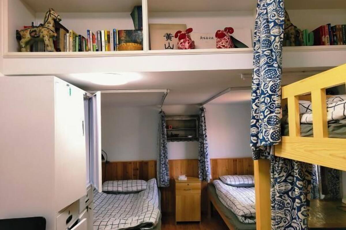 A dormitory room with bunk beds, a bookshelf with books and a horse figurine, and a small wooden cabinet.